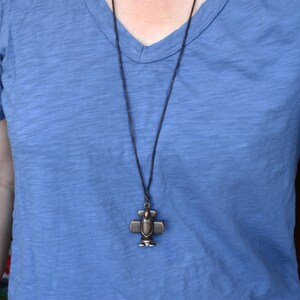 May include: A bronze-colored airplane pendant necklace. The pendant is a detailed miniature airplane design, suspended from a dark cord. The necklace is worn against a blue v-neck shirt.