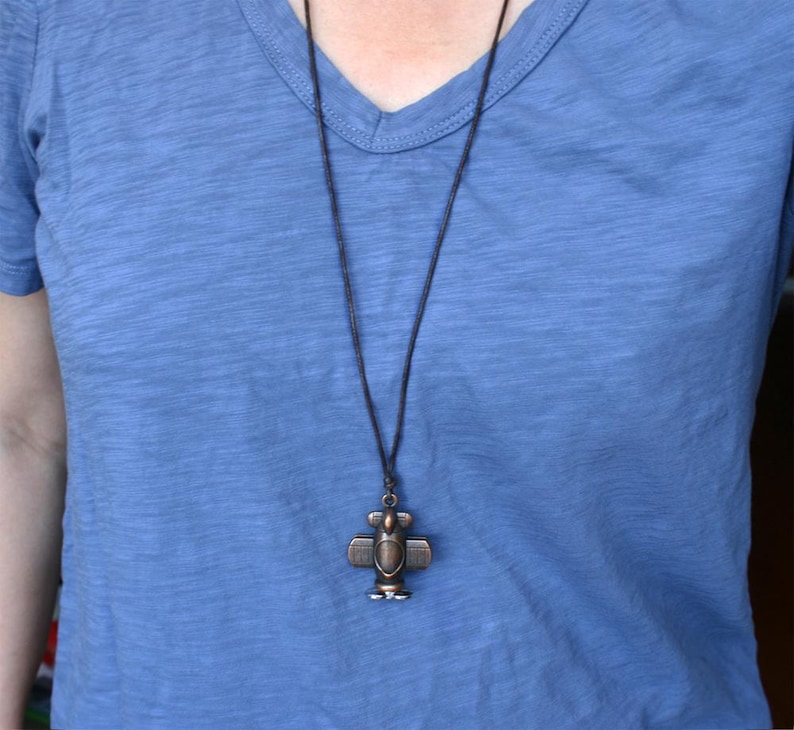 May include: A bronze-colored airplane pendant necklace. The pendant is a detailed miniature airplane design, suspended from a dark cord. The necklace is worn against a blue v-neck shirt.