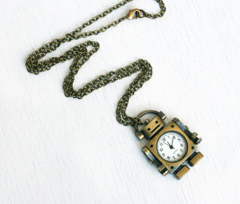 May include: A gold-toned robot-shaped pendant necklace with a clock face. The clock face has black hands and black Roman numeral markings.