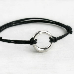 May include: A bracelet featuring a single, polished silver-toned metal ring centered on a thin, black cord. The cord is tied with knots on either side of the ring, creating a simple, circular design that secures the metal component in place.