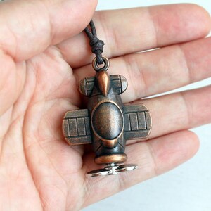 May include: A bronze-colored airplane pendant with a dark brown cord. The pendant features a propeller, wings, and a circular body. The airplane design is detailed and has a vintage aesthetic.