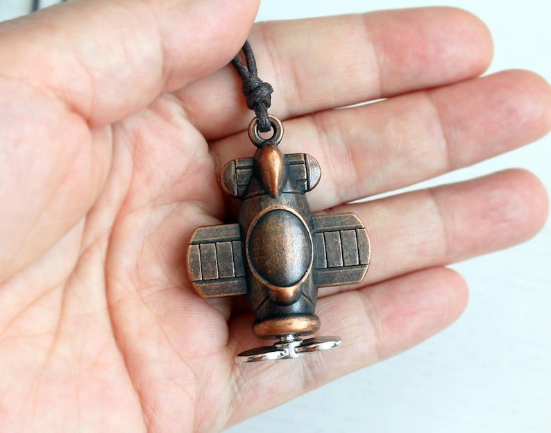 May include: A bronze-colored airplane pendant with a dark brown cord. The pendant features a propeller, wings, and a circular body. The airplane design is detailed and has a vintage aesthetic.