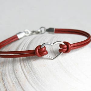 May include: A red leather bracelet with a silver heart charm. The bracelet is made of two strands of leather and has a simple, elegant design.