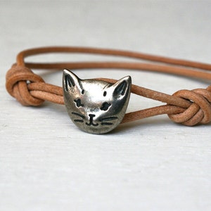 May include: A brown leather bracelet with a silver cat charm. The bracelet is tied with a knot on each end.