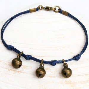 May include: A dark blue cord bracelet featuring three small, round, antique brass-toned jingle bells suspended from the cord. The bracelet is secured with a matching antique brass-toned lobster claw clasp and metal end caps on each side.