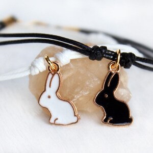May include: Two bracelets featuring gold-toned metal rabbit charms, one filled with white enamel and the other with black enamel. Each charm hangs from a thin cord, with one bracelet on a white cord and the other on a black cord, featuring knots.