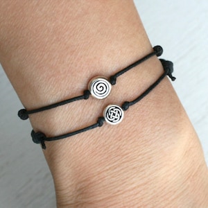 May include: Two black cord bracelets featuring small, round, silver-toned metal beads. One bead displays a spiral design, while the other bead features an intricate, interlaced knot pattern. The bracelets are adjustable with simple sliding knots.