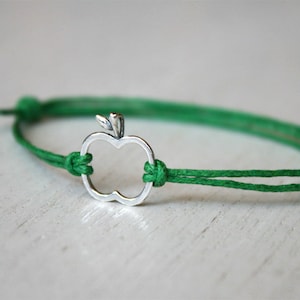 May include: A green string bracelet with a silver apple charm. The apple charm is a simple outline of an apple with a leaf on top.