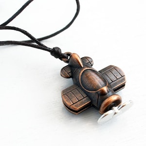 May include: A bronze-colored airplane pendant with a silver propeller hangs from a black cord. The biplane design features detailed wings and a fuselage. The necklace is set against a white background, highlighting the pendant's vintage aesthetic.