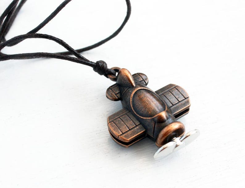 May include: A bronze-colored airplane pendant with a silver propeller hangs from a black cord. The biplane design features detailed wings and a fuselage. The necklace is set against a white background, highlighting the pendant's vintage aesthetic.