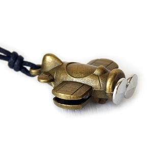 May include: A bronze-colored airplane pendant with a silver propeller is suspended from a black cord. The small, detailed plane has wings and a fuselage. The background is plain white, highlighting the pendant's design.