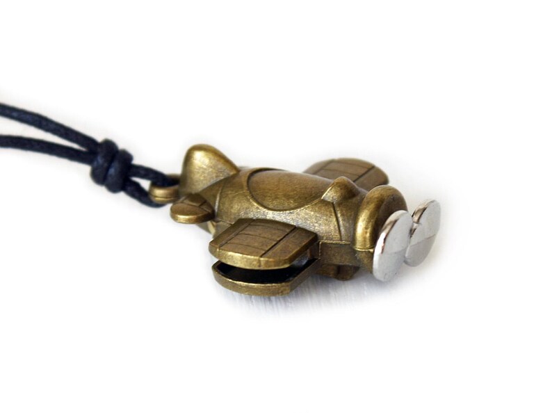 May include: A bronze-colored airplane pendant with a silver propeller is suspended from a black cord. The small, detailed plane has wings and a fuselage. The background is plain white, highlighting the pendant's design.