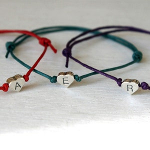 May include: Three personalized heart charm bracelets. Each bracelet features a silver heart charm with a single letter, strung on a cord in red, teal, and purple. The bracelets are knotted for adjustment.