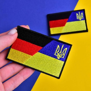 Puede incluir: Dos parches rectangulares bordados. Uno presenta los colores de la bandera alemana (negro, rojo y amarillo) y el escudo de armas ucraniano. El otro parche tiene el mismo diseño. Los parches están sobre un fondo azul y amarillo.