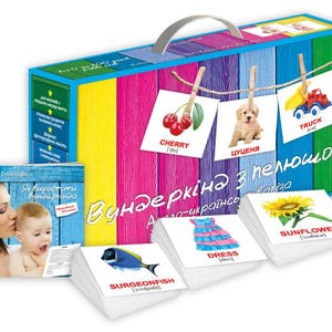 Puede incluir: Un colorido conjunto educativo para niños, con una caja con asa, tarjetas didácticas y un libro. La caja tiene un diseño a rayas con las palabras "Вундеркіна з пелюшок" e imágenes de cereza, cachorro y camión.