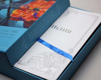 以下が含まれることがあります： 赤い花柄の青い箱で、表紙に「Библия」（聖書）と書かれています。箱が開いており、銀色の模様が施された白い本が現れ、表紙に「Библия」（聖書）と書かれています。