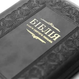 May include: A black book with an embossed decorative border and gold lettering on the cover. The text on the cover reads "БІБЛІЯ" followed by "КНИГИ СВЯТОГО ПИСЬМА" with decorative lines above and below the text. The cover has a matte finish.