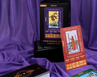 Op de afbeelding: Een set tarotkaarten met een zwarte doos en een paarse achtergrond. De doos heeft de tekst "ORNER" en "НАВЧАЛЬНІ ТАРИ РАЙДЕРА-УЕЙТА" erop. De kaarten zijn in verschillende kleuren, waaronder rood, groen en blauw. De kaarten hebben afbeeldingen van mensen, dieren en objecten. De kaarten worden gebruikt voor waarzeggerij en het voorspellen van de toekomst.