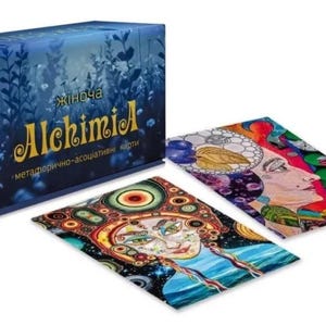 May include: A dark blue box set of "AlchimiA" cards with a floral design. Two colourful tarot cards are displayed, featuring artistic illustrations of faces and abstract patterns. The text on the box is in a foreign language.