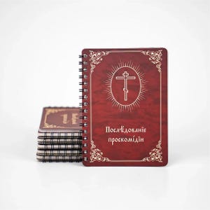 May include: A stack of spiral-bound notebooks with a burgundy cover. The front notebook features a white cross within a circle and text in a script font. The cover has ornate gold corner designs.