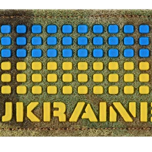Puede incluir: Un parche rectangular con la palabra "UKRAINE" en letras amarillas. La parte superior presenta cuadrados azules y la inferior cuadrados amarillos, asemejándose a una bandera. El parche tiene un borde verde tipo camuflaje.