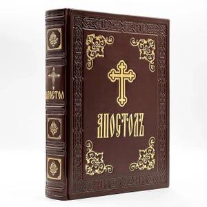 May include: A brown leather-bound book with gold embossed accents and lettering. The cover features a gold cross and the word "Apostol" in gold lettering. The spine has decorative gold squares and a cross.