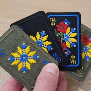 以下が含まれることがあります： ウクライナのフォークアートデザインの刺繍パッチ。パッチは、黒とオリーブグリーンの背景に、黄色、青、赤の花柄と幾何学模様が特徴です。パッチは角が丸い長方形です。