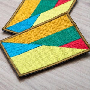 May include: Two rectangular embroidered patches featuring a geometric pattern of yellow, green, blue, and red diagonal segments. Each patch is finished with a dark brown stitched border, creating a clean edge around the colorful fabric design.