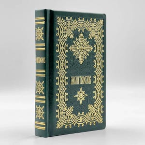 May include: A dark green book with gold embossed decorative patterns and text. The book's cover features ornate designs and the title "MOLITVOSLOV" in gold lettering. The spine also has gold accents. The book is standing upright.