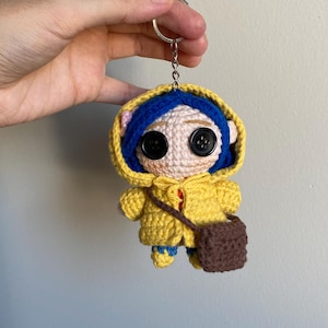 May include: A handmade crochet keychain featuring a doll character with button eyes, blue hair, and a yellow hooded coat. The doll has a small brown satchel and blue shoes. The keychain is attached to a silver ring.