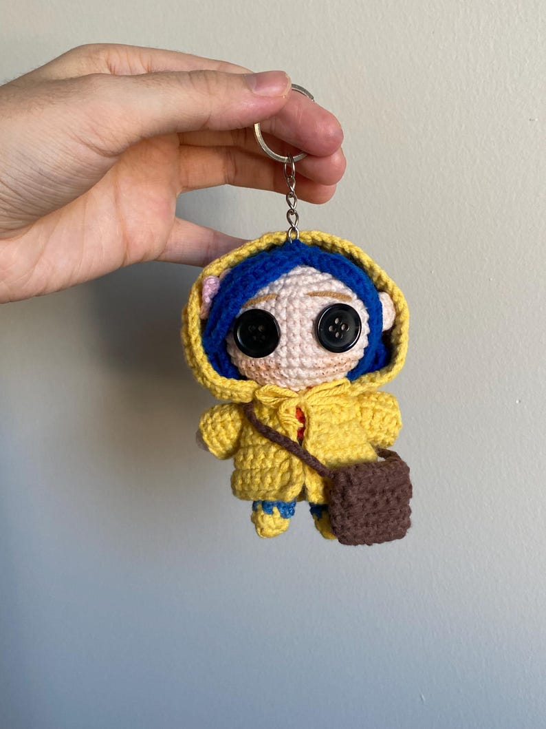 May include: A handmade crochet keychain featuring a doll character with button eyes, blue hair, and a yellow hooded coat. The doll has a small brown satchel and blue shoes. The keychain is attached to a silver ring.