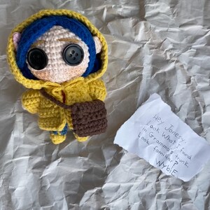 May include: A handmade crochet doll of a character with button eyes, wearing a yellow hooded jacket, blue hair, and a brown purse. A handwritten note reads "Hey Jonesy. Look What I found in Gramma's trunk. Look familiar? Wybie."