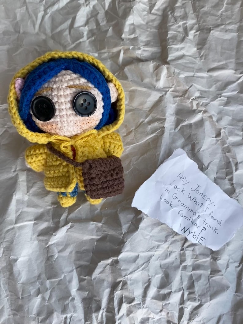 May include: A handmade crochet doll of a character with button eyes, wearing a yellow hooded jacket, blue hair, and a brown purse. A handwritten note reads "Hey Jonesy. Look What I found in Gramma's trunk. Look familiar? Wybie."