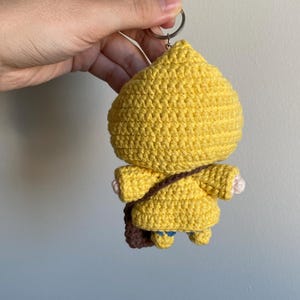 May include: A handmade yellow crochet keychain in the shape of a character wearing a yellow coat and a brown satchel. The keychain is attached to a silver metal ring. The character has small blue accents on the feet and white hands.