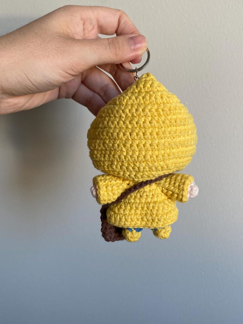 May include: A handmade yellow crochet keychain in the shape of a character wearing a yellow coat and a brown satchel. The keychain is attached to a silver metal ring. The character has small blue accents on the feet and white hands.