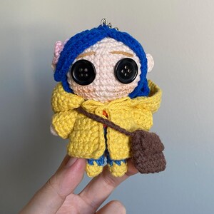 May include: Crocheted doll featuring a character with blue hair, button eyes, and a yellow coat. The doll has a small brown satchel and is wearing blue pants. A pink flower adorns the character's hair. The doll is a handmade item.