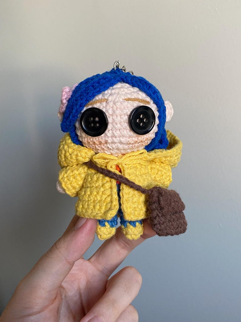 May include: Crocheted doll featuring a character with blue hair, button eyes, and a yellow coat. The doll has a small brown satchel and is wearing blue pants. A pink flower adorns the character's hair. The doll is a handmade item.