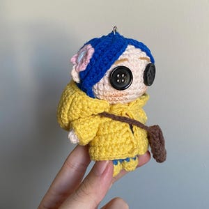 May include: A handmade crochet doll with a yellow hooded jacket, blue hair, and large button eyes. The doll has a small brown satchel and a pink flower on its head. The doll is held by a hand.