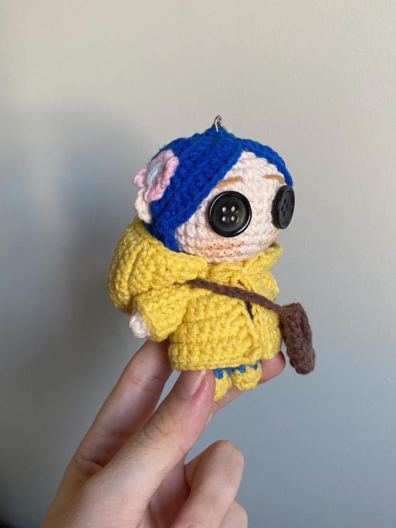 May include: A handmade crochet doll with a yellow hooded jacket, blue hair, and large button eyes. The doll has a small brown satchel and a pink flower on its head. The doll is held by a hand.
