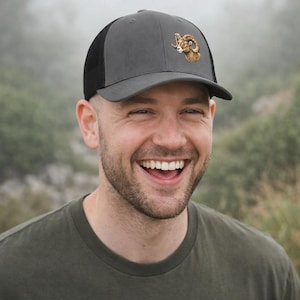 May include: Gray and black trucker hat featuring a detailed illustration of a ram's head on the front. The hat has a curved brim and a mesh back for breathability. A stylish accessory for outdoor enthusiasts.