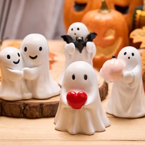 Könnte beinhalten: Eine Sammlung weißer Keramik-Geisterfiguren mit schwarzen Punktaugen und lächelnden Gesichtern. Ein Geist hält ein rotes Herz, ein anderer einen kleinen Kürbis und ein weiterer eine Fledermaus. Geschnitzte Kürbisse und Herbstblätter im Hintergrund.