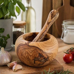 Op de afbeelding: Een houten vijzel en stamper set met een natuurlijk houtnerfpatroon. De vijzel is rond en de stamper is lang en cilindrisch. De set staat op een houten snijplank, met knoflook, tomaten en kruiden in de buurt. Een keukenhulp.