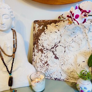 May include: A white ceramic Buddha statue with a brown wooden beaded necklace. The statue is sitting on a table with a white ceramic candle holder and a white and brown ceramic tile with a floral design. A white orchid with pink and purple accents is in the background.
