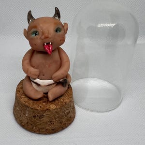 May include: A small, brown, clay figurine of a devil baby with horns, wearing a nappy, sitting on a cork base. The figurine is under a glass dome.