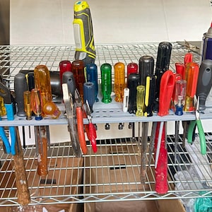 May include: A gray plastic tool organizer rack holding a variety of hand tools including screwdrivers with colorful handles, pliers, a yellow and black power tool, and a blue and yellow can of WD-40 Dry Lube, all arranged on a wire shelving unit.