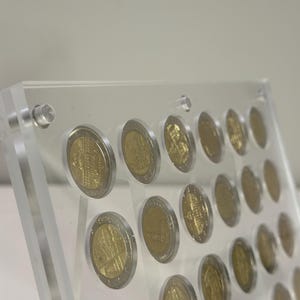 Puede incluir: Una vitrina transparente de acrílico que contiene varias monedas de euro doradas y plateadas. Las monedas están dispuestas en filas y columnas, mostrando sus diseños detallados. La vitrina está asegurada con cierres transparentes.