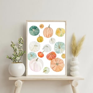 Puede incluir: Una impresión en acuarela enmarcada que presenta una colección de calabazas de varios tamaños y colores. Las calabazas varían de suaves pasteles a naranjas y verdes vibrantes, creando un tema otoñal. La obra de arte se muestra sobre un fondo blanco.