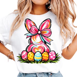 May include: White t-shirt featuring a colorful Easter bunny design. The bunny is pink with a large bow and surrounded by decorated Easter eggs in various patterns and colors. The design is set against a backdrop of green grass.