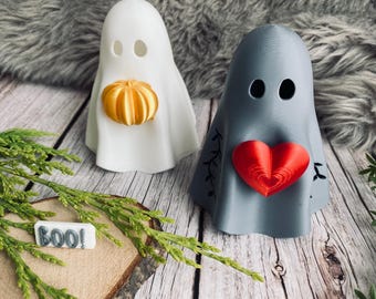 Könnte beinhalten: Zwei 3D-gedruckte Geisterfiguren, einer weiß mit einem Kürbis, der andere grau mit einem roten Herzen. Das Wort "BOO!" steht auf einem kleinen Schild. Halloween-Dekorationen.
