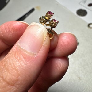 May include: A pair of small stud earrings featuring round, faceted pink-red gemstones set in gold-toned metal prongs. The earrings have a post-back design with visible metal butterfly clutch backings attached to the posts behind the stone settings.